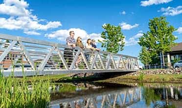 Four friends stand on the bridge and lean over the railing, looking out at the water on a bright, sunny day.